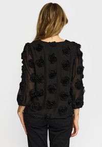 Sort gennemsigtig bluse med tredimensionelle sorte roseudsmykninger over det hele. Lange ærmer med elastiske manchetter. Glat tekstur.