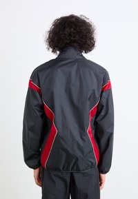 Black windbreaker jacket with red and white accents, featuring a high collar and elasticized cuffs, smooth texture, and a relaxed fit.