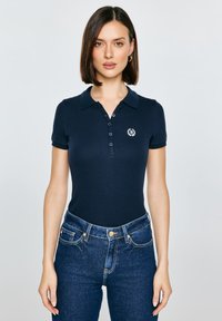 Námořnická polokošile s límečkem, opatřená šesti knoflíky a vyšitým logem. Kombinováno s modrými džínami s vysokým pasem.