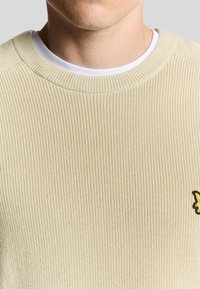 Beige ribbet striktrøje med rund halsudskæring. Bemærkelsesværdig gul og sort logo på venstre bryst, lagdelt over en hvid skjortekrave.