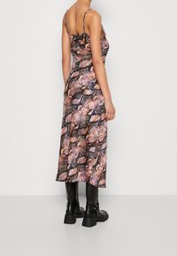 Femme portant une robe midi sans manches à imprimé serpent rose et noir, avec des bottes à talons noires, se tenant devant un fond clair.