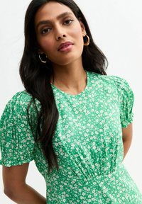 Vestido floral verde con mangas cortas abullonadas, tejido ligero, detalles fruncidos en la cintura y un escote redondo. Patrones florales blancos en todo el vestido.