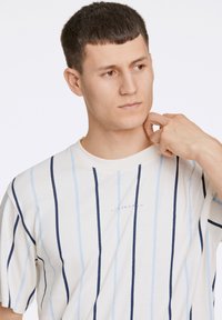 Witte t-shirt met verticale marineblauwe en lichtblauwe strepen, ronde hals, korte mouwen en logo in het midden op de borst. Zachte stoftextuur.