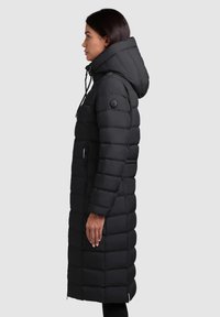 Longue doudoune noire avec capuche, présentant un design matelassé, une fermeture à cordon de serrage et une poche zippée sur le côté.