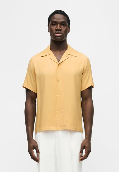 Άνδρας που στέκεται με το πρόσωπο προς τα εμπρός, φοράει ένα κοντομάνικο πουκάμισο μουσταρδί με κουμπιά και λευκό παντελόνι, μπροστά σε ένα απλό φόντο.