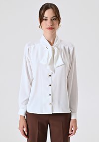 Witte blouse met een strikje kraag, lange mouwen, knoopsluiting aan de voorkant en een subtiele glans. Bevat lichtgekleurde knopen en een gladde stofstructuur.