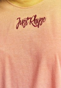 Lys oransje T-skjorte med rund hals, med maroon brodert tekst "JustRhyse." Myk bomullsone med lett tekstur.