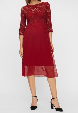 Kvinde iført en knælængde rød blondekjole med trekvartærmer og sorte ankelremssandaler, stående mod en ensfarvet baggrund.