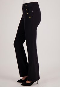 Zwarte broek met uitlopende pijpen van rekbaar materiaal, met hoge taille, drie grote gouden knopen en zijzakken. Gladde textuur.