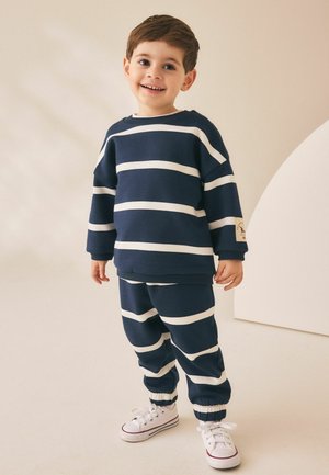 Junger Junge lächelt, trägt einen marineblau-weiß gestreiften Pullover und passende Hose mit weißen Turnschuhen, steht drinnen auf beigem Boden.