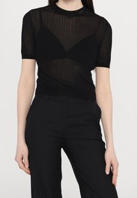 Czarny krótki top z przezroczystej, teksturowanej tkaniny z pionowymi paskami i zaokrąglonym dekoltem, noszony na czarnym topie na ramiączkach.