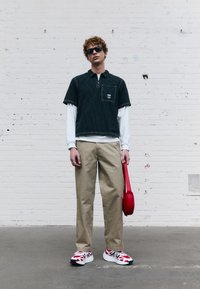 Camicia a maniche corte verde scuro con colletto e motivo a righe sopra una maglietta a maniche lunghe bianca, abbinata a pantaloni beige e una borsa rossa. Sneakers rosse e bianche.