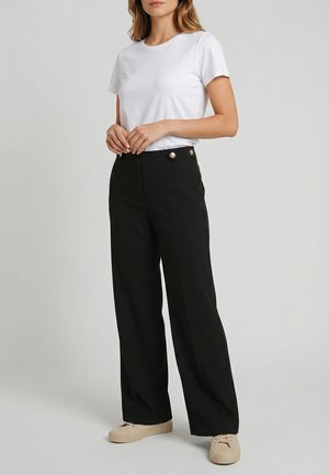 Donna che indossa una maglietta bianca semplice a maniche corte, pantaloni neri a gamba larga a vita alta con bottoni dorati e scarpe casual beige, in piedi su uno sfondo semplice.