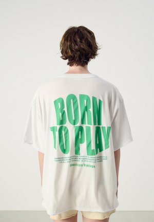 Osoba o krótkich brązowych włosach stoi tyłem, ubrana w białą, dużą koszulkę z dużym zielonym napisem "BORN TO PLAY" na plecach.