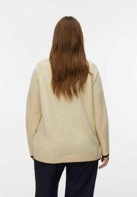 Femme aux longs cheveux bruns portant un pull beige et un pantalon sombre, se tenant dos à la caméra devant un fond blanc uni.