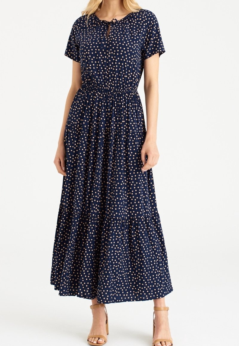Femme portant une robe maxi bleu marine à manches courtes avec des pois rose clair, associée à des talons ouverts beige.