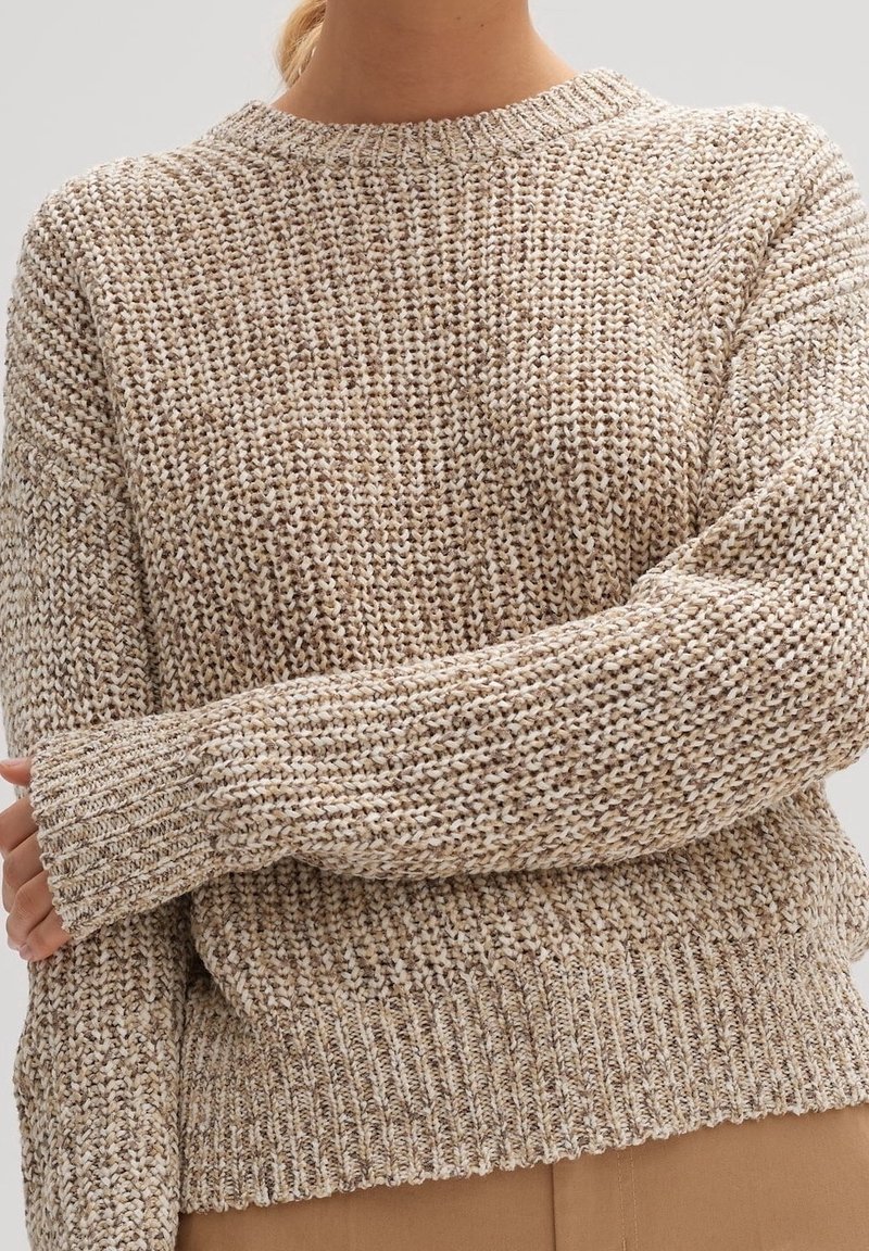 Beżowy sweter dziany z wzorem teksturowym, okrągłym dekoltem i ściągaczami. Materiał wydaje się miękki i gruby, zapewniając ciepło oraz komfort.