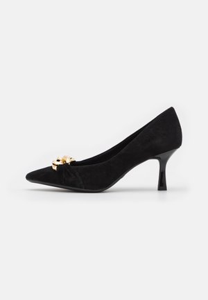 Zwarte suède kittenheel pump met spitse neus en gouden kettingversiering op een witte achtergrond.
