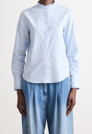 Person iført en lyseblå, langærmet skjorte med knapper, flæsekant ved kraven og manchetterne, kombineret med løst siddende blå denimbukser.