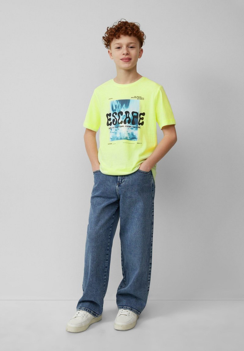 Neon gele katoenen T-shirt met een "ESCAPE" graphic en blauwe waterpatronen. Gecombineerd met een losse fit blauwe denim jeans en witte sneakers.