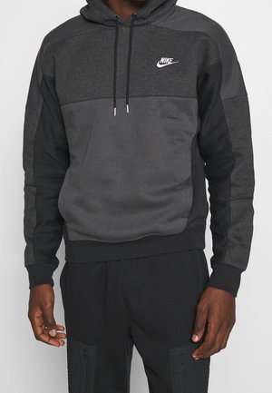 Man draagt een donkergrijze en zwarte Nike-hoodie met logo op de borst en bijpassende zwarte broek, staand met ontspannen handen langs het lichaam.
