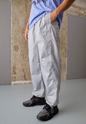 Person iført lysegrå Nike Air bukser med elastiske manchetter og sorte sneakers, stående indendørs mod en tekstureret væg.