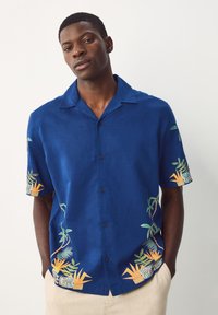 Chemise à manches courtes en lin bleu avec broderie tropicale en orange et vert, dotée d'un col et de boutons sur le devant.