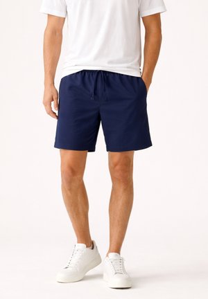 Uomo che indossa pantaloncini con coulisse blu navy, maglietta bianca a girocollo e sneakers bianche, in piedi con una mano in tasca.