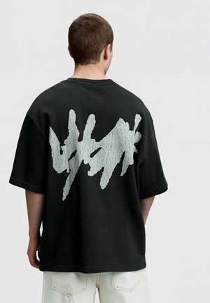 Junger Mann trägt ein schwarzes oversized T-Shirt mit einem großen abstrakten weißen Farbspritzer-Design auf dem Rücken, kombiniert mit hellen Hosen.