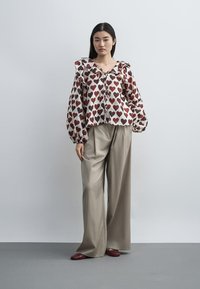 Femme portant un chemisier blanc à motifs de cœurs rouges, un pantalon large beige et des chaussures rouges, debout devant un fond uni gris clair.