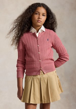 Meisje met lang krullend haar dat een roze kabelgebreid vest draagt over een wit overhemd met kraag en een beige geplooide rok, staand tegen een effen achtergrond.