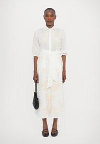 White linen dress with a soft yellow floral pattern, short puff sleeves, a collar, and a tied waist. Black handbag and shoes accent the outfit.