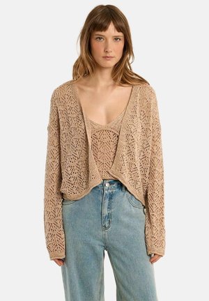 Kvinde iført beige åbenstrikket cardigan og matchende top med lyseblå højtaljede jeans, stående foran en ensfarvet baggrund.