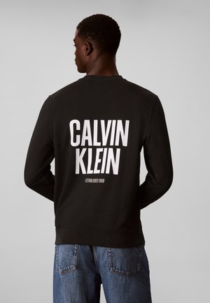 Mężczyzna widziany od tyłu w czarnej bluzie z dużym białym tekstem "Calvin Klein" i niebieskich dżinsach na jednolitym szarym tle.