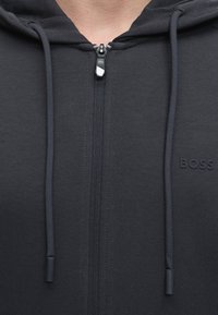 Gros plan d'un sweat à capuche noir zippé avec cordons et un logo discret "BOSS" sur la poitrine.