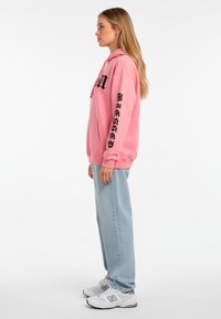 Felpa con cappuccio rosa con testo nero sulle maniche, abbinata a pantaloni blu chiaro e sneakers bianche con accenti grigi, mostrata di lato.