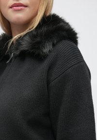 Pulover negru cu un design texturat, cu dungi și o garnitură din blană artificială la glugă. Prezintă un fermoar la gât și o croială lejeră.