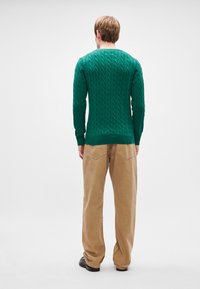 Πράσινο πουλόβερ με πλέξη καλωδίου και στρογγυλή λαιμόκοψη, μακρυά μανίκια και ραμμένες μανσέτες, σε συνδυασμό με μπεζ παντελόνι σε φαρδύ κόψιμο.