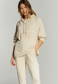 Beige huppari ja yhdisteet pehmeästä kankaasta. Hupparissa on kiristysnaru ja hienovarainen logomuotoilu. Rentoa istuvuutta, jossa on sivutaskut.