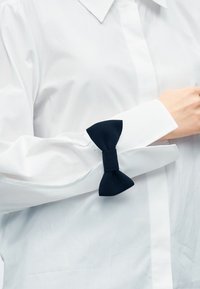 Λευκό πουκάμισο με υφή, που διαθέτει σκούρο μπλε φιόγκο στο μανίκι. Ο γιακάς είναι μυτερός και τα μανίκια είναι απαλά συστραμμένα.