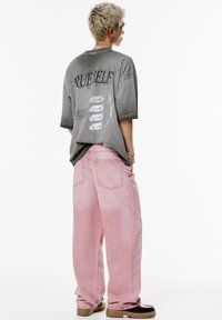 Person med kort blondt hår iført en oversized grå grafisk t-shirt, lyserøde brede bukser og brune plateausko, stående foran en hvid baggrund.