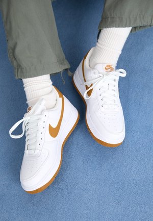 Personne portant des baskets blanches Nike Air Force 1 avec un swoosh et des semelles marron, associées à des chaussettes blanches et un pantalon vert sur un tapis bleu.