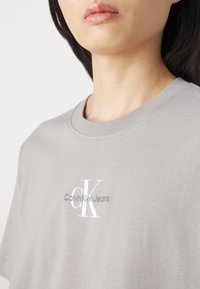 Pelēks kokvilnas T-krekls ar apaļu apkakli un īsām piedurknēm. Uz priekšpuses izšūts "Calvin Klein Jeans" logo baltā un gaiši pelēkā krāsā.