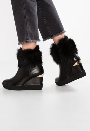 Czarne skórzane botki na klinie z czarnym futrzanym wykończeniem i złotym detalem na obcasie, noszone z poszarpanymi czarnymi dżinsami.