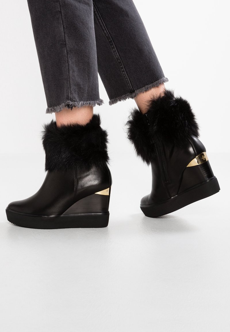 Bottines compensées en cuir noir avec garniture en fourrure noire et détail doré au talon, portées avec un jean noir effiloché.