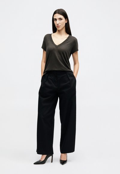 Femme aux longs cheveux noirs portant un T-shirt à col en V sombre, un pantalon large noir et des chaussures à talons noires, debout les mains dans les poches.