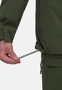 Giacca verde traspirante con una texture liscia, dotata di lacci regolabili all'orlo e tasca con zip sul lato inferiore.