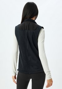 Gilet in fleece nero con pannello spalla nero liscio, colletto alto e orlo regolabile. Indossato sopra una maglietta bianca a maniche lunghe.