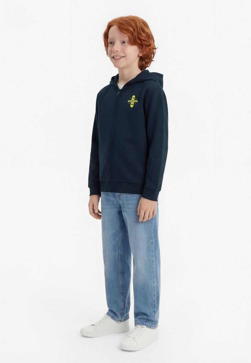 Garçon aux cheveux roux debout, portant un sweat à capuche zippé bleu marine avec un logo jaune, un jean bleu clair et des baskets blanches, souriant légèrement sur le côté.