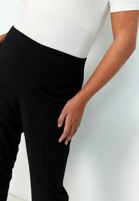 Black, high-waisted pants with smooth fabric and a tailored fit. Close-up shows hand resting on the leg, highlighting sleek design.